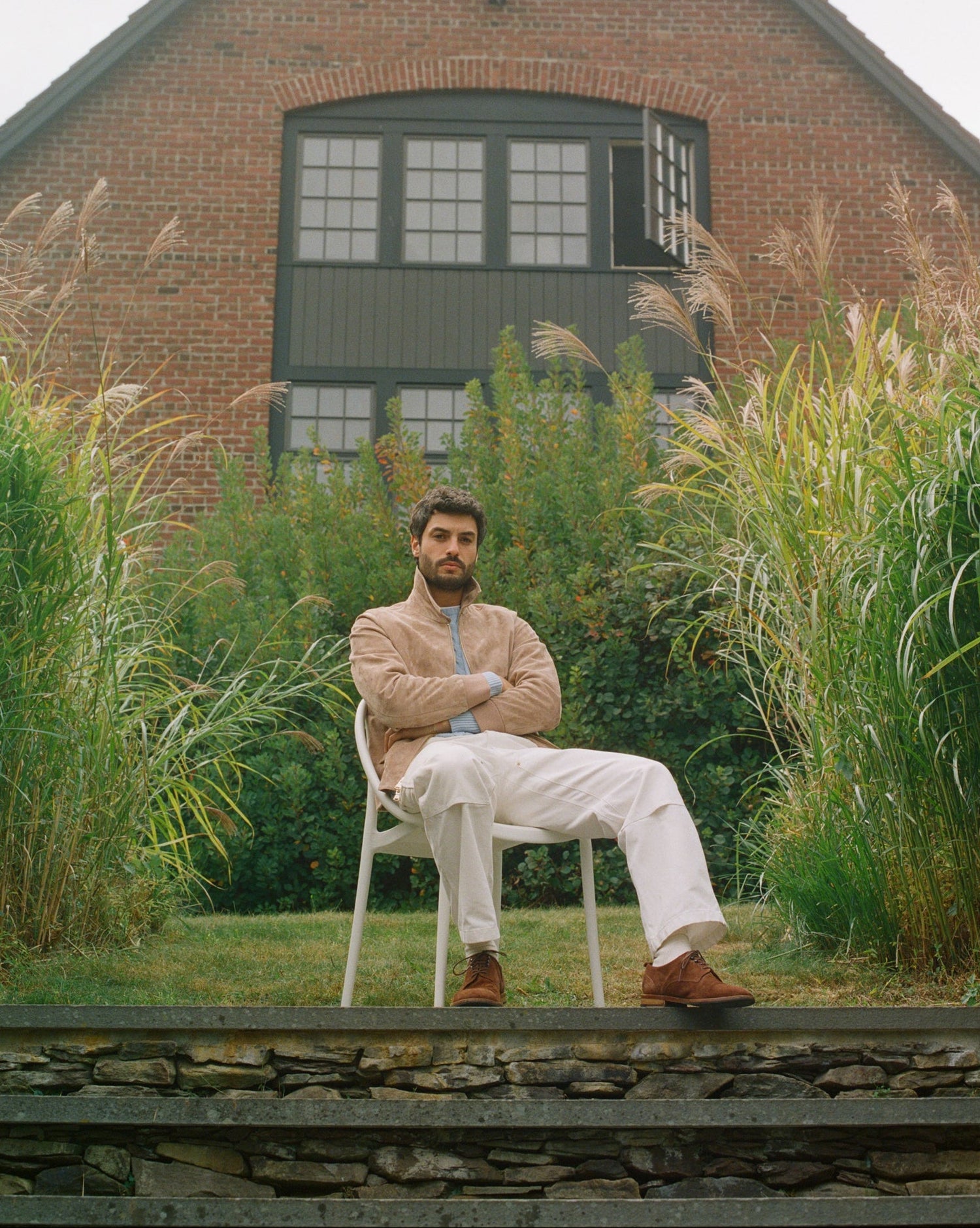 Man sitting on a chair in front of a brick building with tall grasses around.